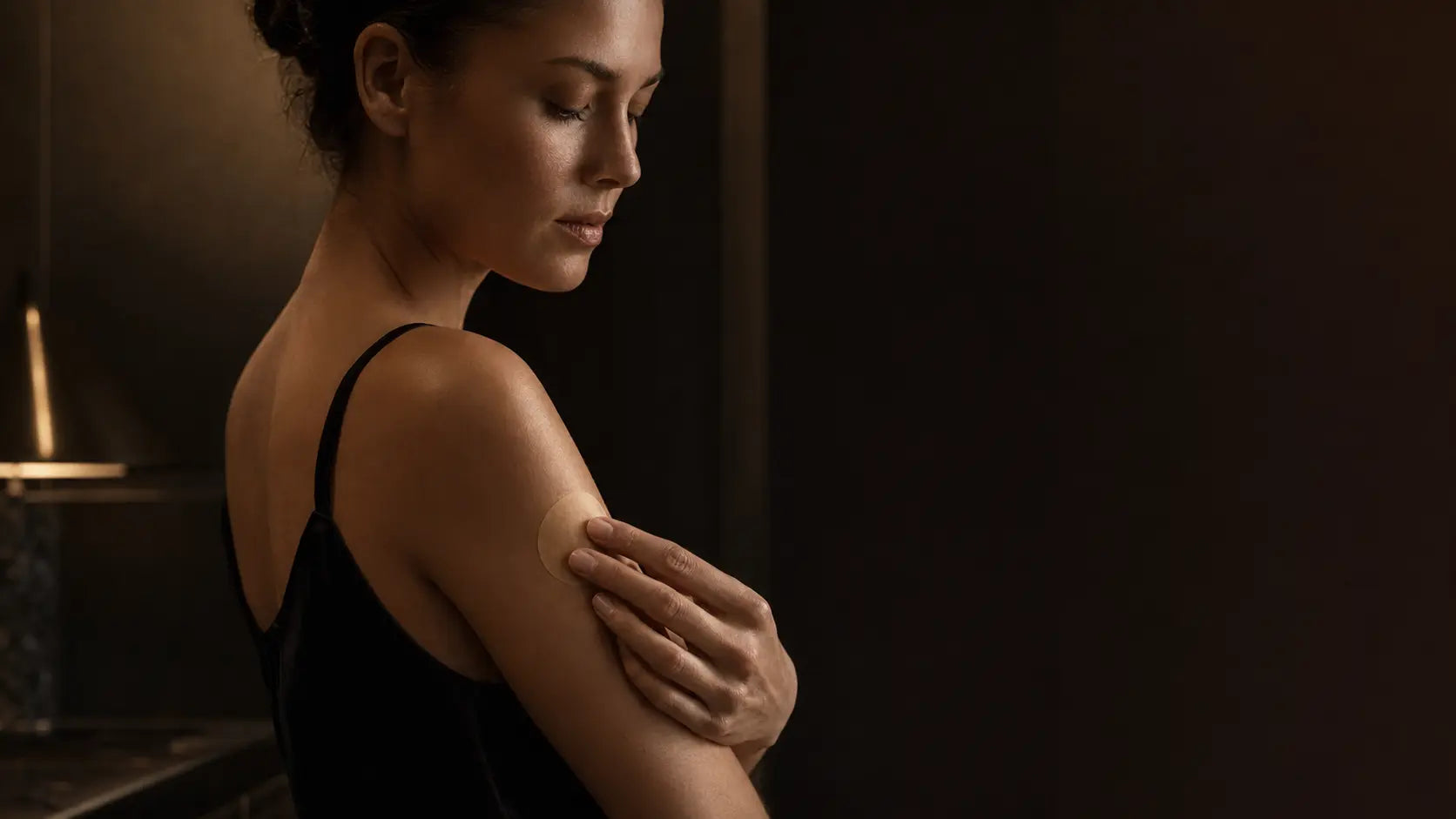 A woman applying a discreet circular skincare patch to her upper arm in warm atelier light
